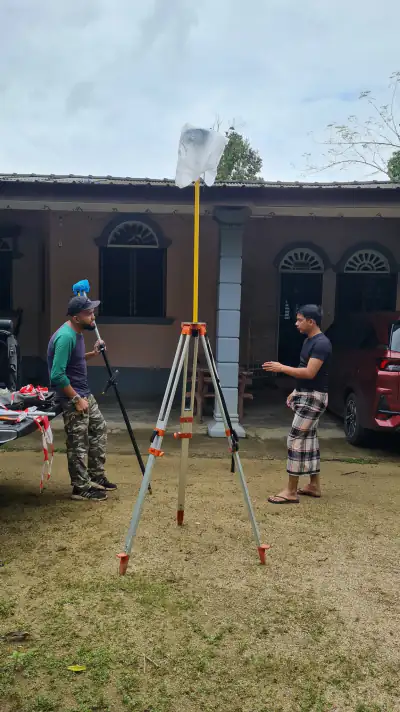 Tripod dan rod ukur GNSS di halaman rumah kampung; seorang anggota pasukan berbual dengan seorang lelaki, sebuah kereta di sisi.
