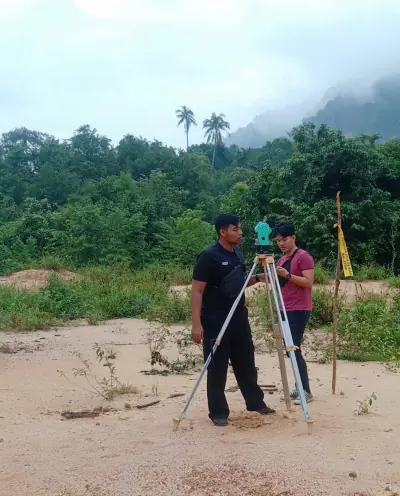 Dua anggota pasukan mengendalikan total station di atas tripod di tapak lapang, dengan latar belakang bukit berkabus.