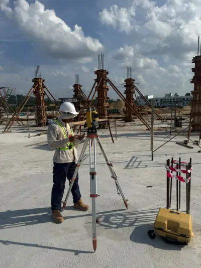 Seorang anggota pasukan bertopi keledar mengendalikan total station di tapak pembinaan dengan acuan tiang konkrit di latar.