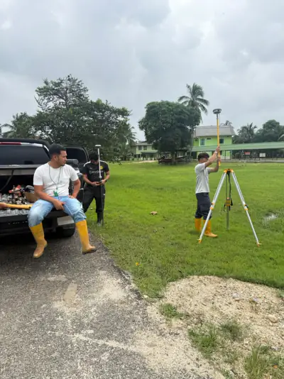 Pasukan Abe memasang tripod dan rover GNSS di padang berumput berhampiran pacuan empat roda, bangunan di latar.