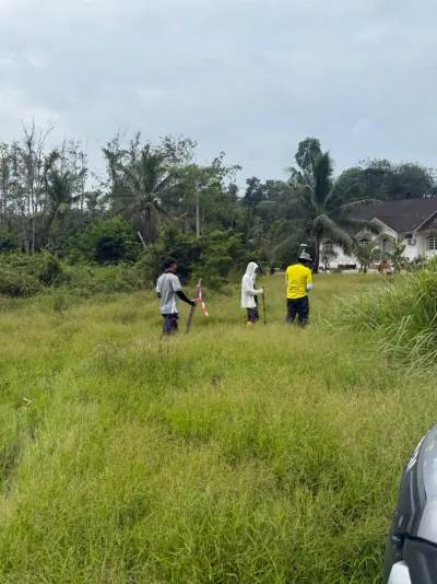 Tiga pekerja meredah tanah berumput tinggi membawa rod ukur dan tiang penanda.