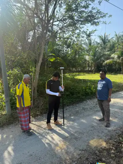 Seorang anggota pasukan mengambil bacaan rover GNSS di jalan kampung sambil dua orang melihat.